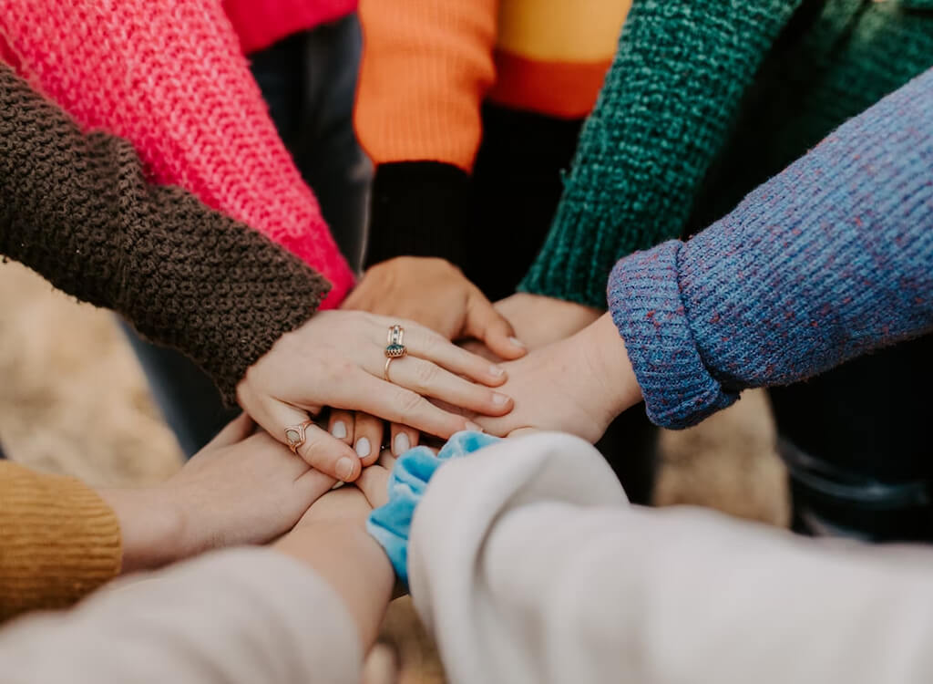 Image of 7 people placing their hands together in a circle as a sign of teamwork and community